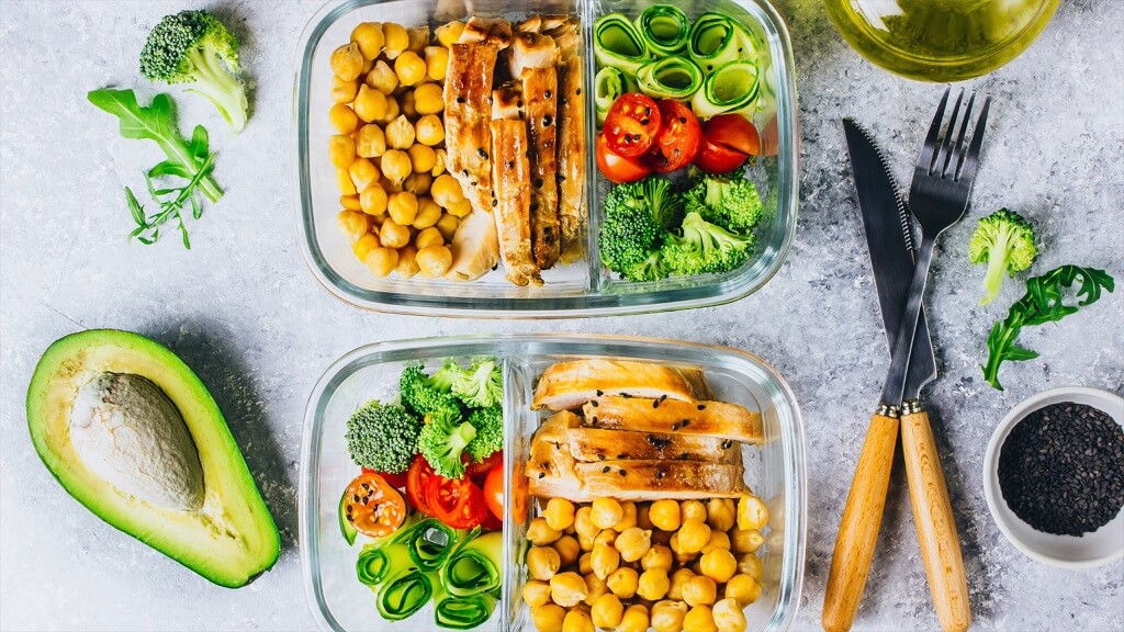 Two glass meal prep containers filled with chickpeas, grilled chicken, and assorted vegetables on a countertop with avocado and utensils